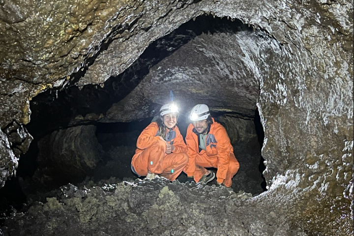 Two people in orange suits and helmets in a rocky cave with headlamps illuminating the area.