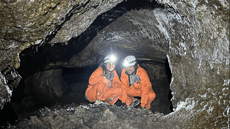 Two people in orange suits and helmets in a rocky cave with headlamps illuminating the area.