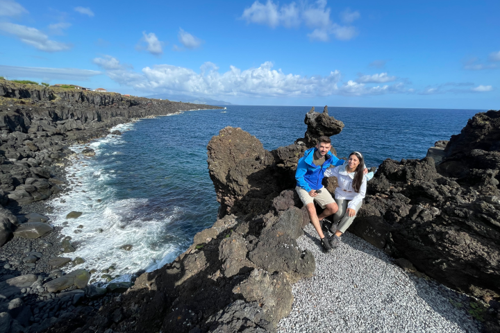 Two people sitting on rocky coast overlooking the ocean with a clear blue sky.