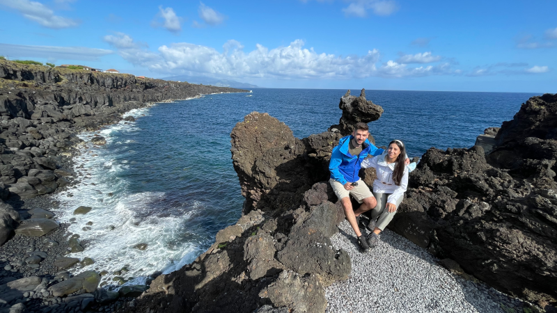 Two people sitting on rocky coast overlooking the ocean with a clear blue sky.