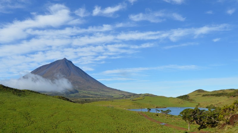 Mountain with clouds, green hills, and small lake under blue sky.