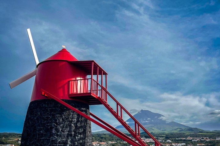Red windmill with a stone base against a mountainous backdrop and a cloudy sky.