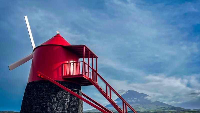 Red windmill with a stone base against a mountainous backdrop and a cloudy sky.