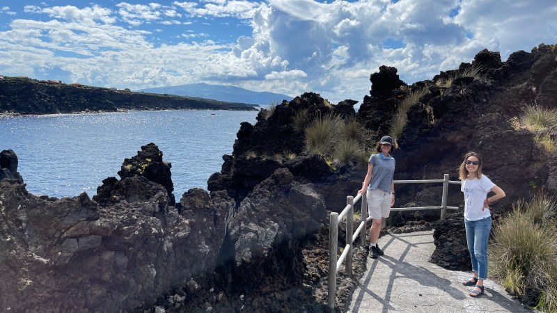 Two people on a rocky coastal path overlooking the sea under a partly cloudy sky.