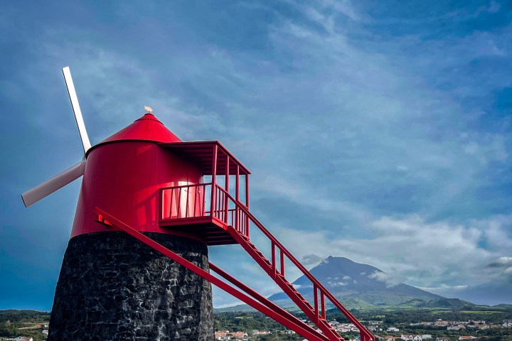 Red windmill on stone base against blue sky, with mountain and village in the background.