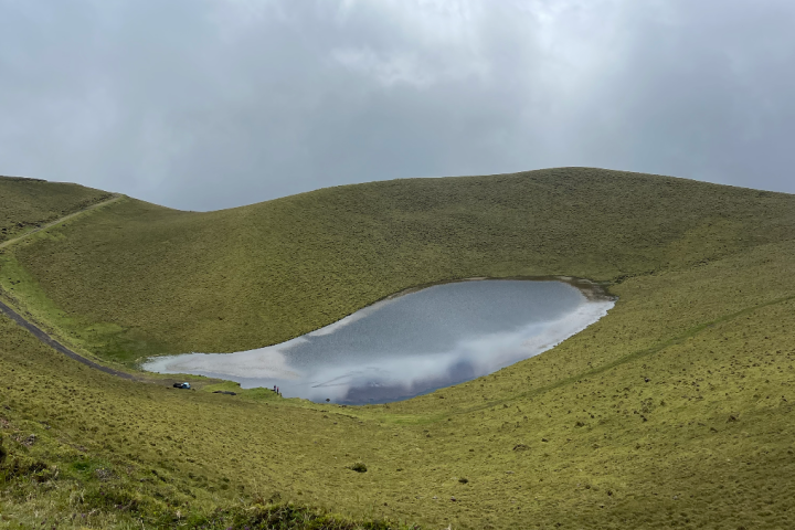 Small lake surrounded by grassy hills under cloudy sky.