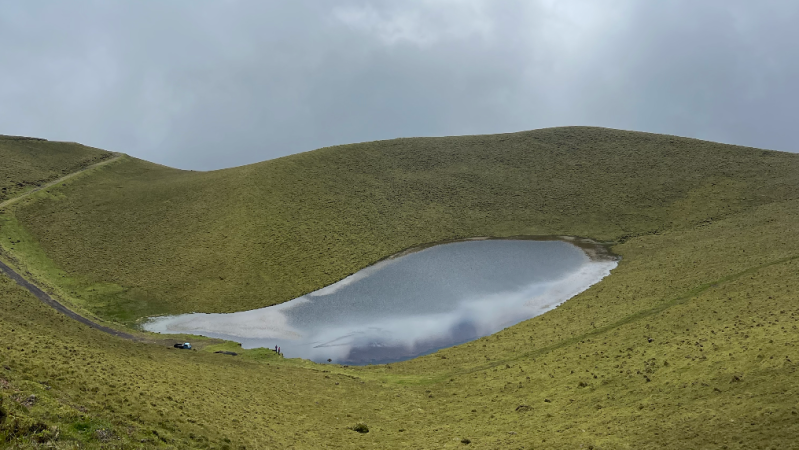 Small lake surrounded by grassy hills under cloudy sky.