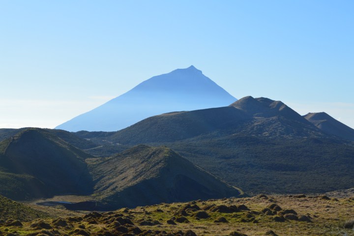 A distant mountain peak with rolling hills and grass in the foreground under a clear blue sky.