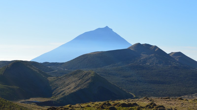 A distant mountain peak with rolling hills and grass in the foreground under a clear blue sky.