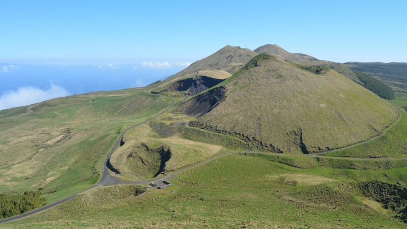 Green hills with winding road under clear blue sky and distant ocean view.