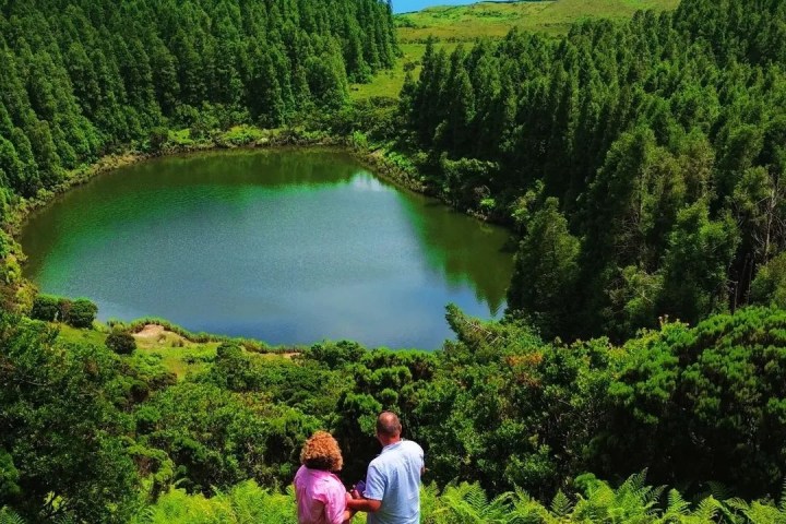 Couple stands on hillside overlooking lush green forest and a round lake.