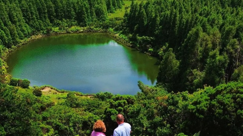 Couple stands on hillside overlooking lush green forest and a round lake.