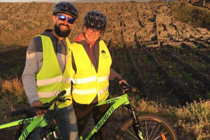 Two cyclists in yellow vests pose with bikes in front of a mountain.