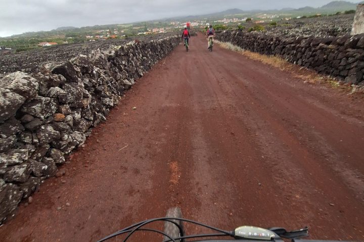 Cyclists on a red dirt road with stone walls and overcast sky.
