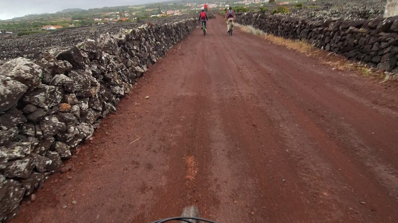 Cyclists on a red dirt road with stone walls and overcast sky.