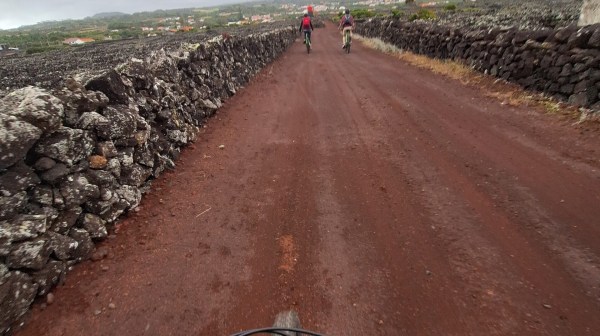 View from bicycle handlebars on a dirt road with two cyclists ahead, cloudy sky.