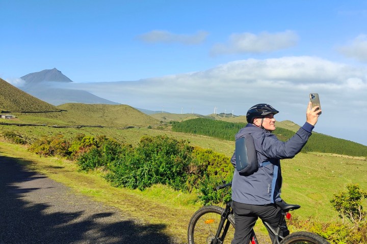 Cyclist taking a selfie on a scenic road with mountains and blue sky in the background.