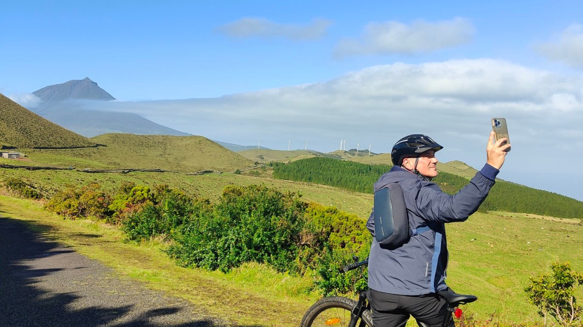 Cyclist taking a selfie on a scenic road with mountains and blue sky in the background.