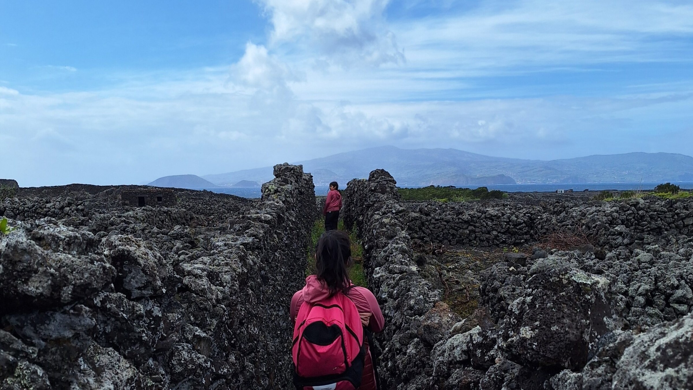 Two people walk between stone walls under a cloudy blue sky with mountains in the distance.