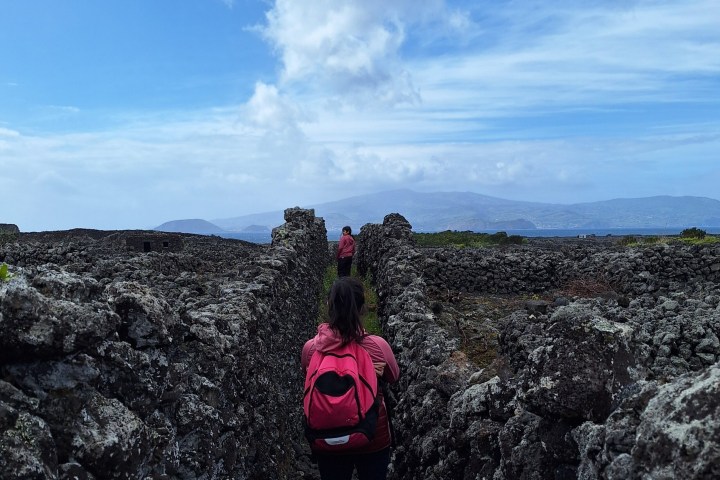 Two people walk between stone walls under a cloudy blue sky with mountains in the distance.