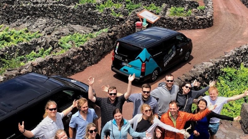 Group of 14 people posing cheerfully next to two vans on a stone path through green fields.