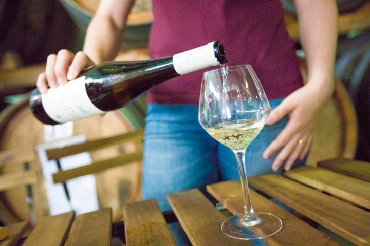 Person pouring white wine into a glass on a wooden table in a cellar.