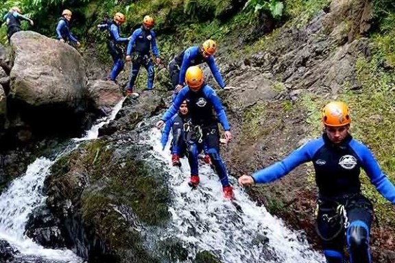 Group of people canyoning with helmets and wetsuits, descending a rocky stream in a forest.