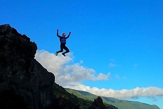 Person jumping off a rocky cliff against blue sky and distant mountains.
