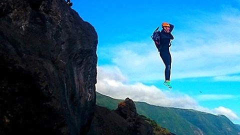 Two people in helmets cliff jumping into the ocean with a mountainous coastline in the background.