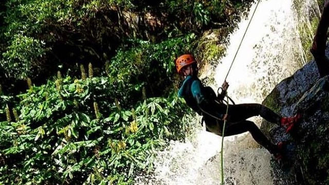 Person rappelling down a waterfall with lush greenery around, wearing a helmet and harness.
