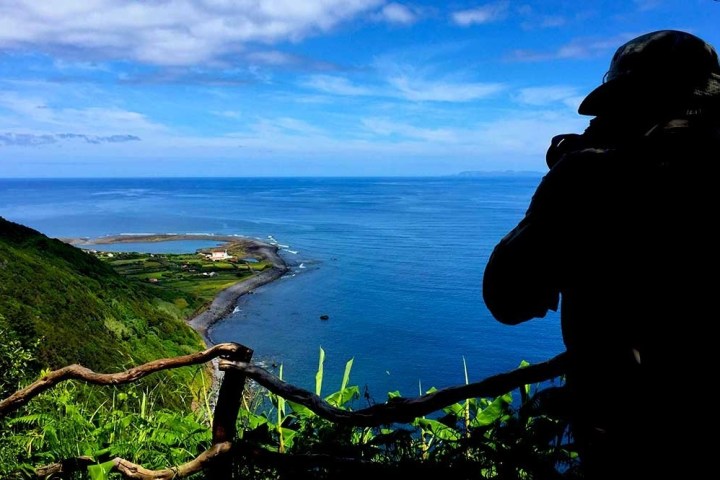 Silhouette of person with camera overlooking coastal landscape and ocean under blue sky.