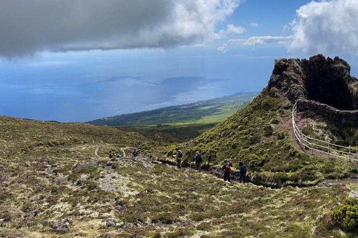 Hikers on a mountainous trail with ocean and clouds in the background.