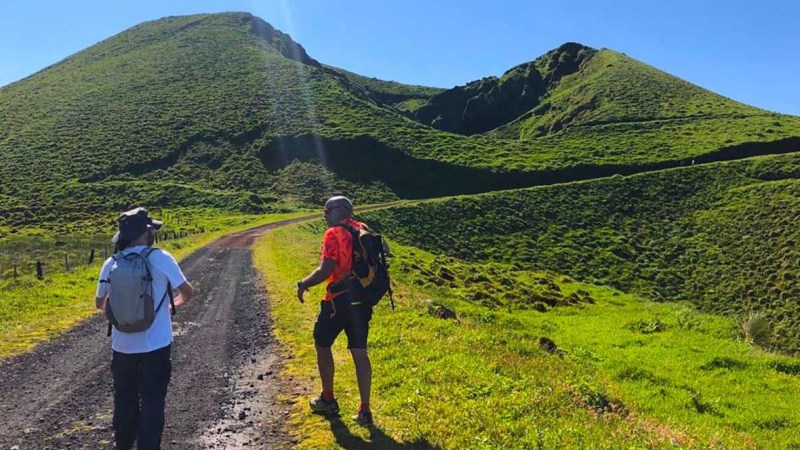Two hikers on a dirt path with green hills in the background under a clear blue sky.