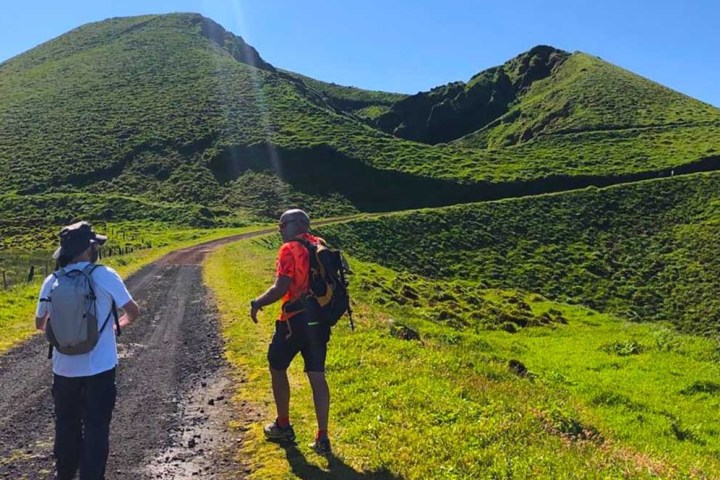 Two hikers walking on a grassy trail toward green hills under a clear blue sky.