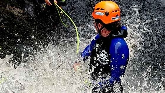 Two people wearing helmets and wetsuits canyoning in rushing water with a rope.