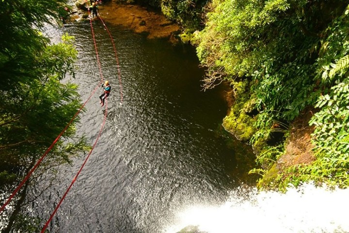 Person ziplining over a river near a waterfall, surrounded by lush greenery.