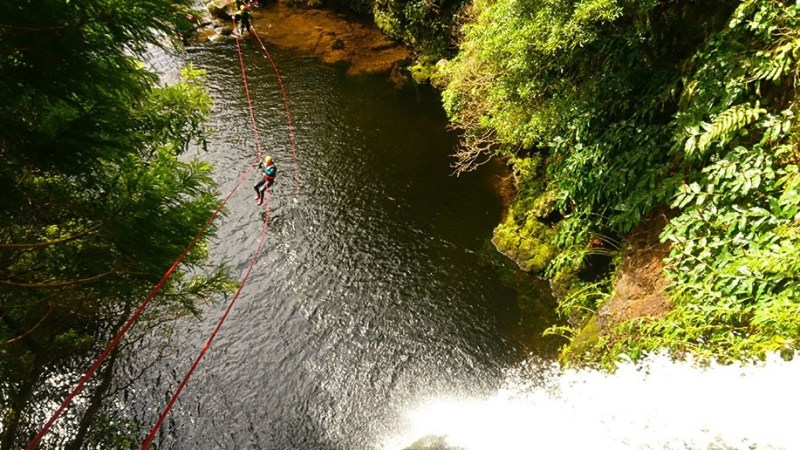 Person ziplining over a river near a waterfall, surrounded by lush greenery.