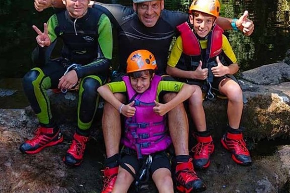Group of four people wearing helmets and wetsuits sitting on rocks near water, smiling and giving thumbs up.