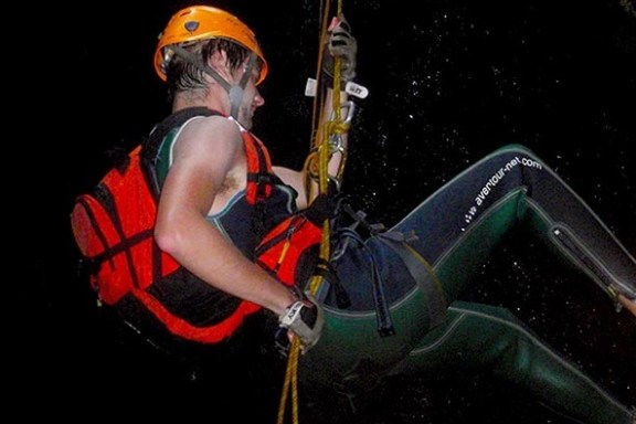Person rappelling in wetsuit, helmet, and harness against a dark background.