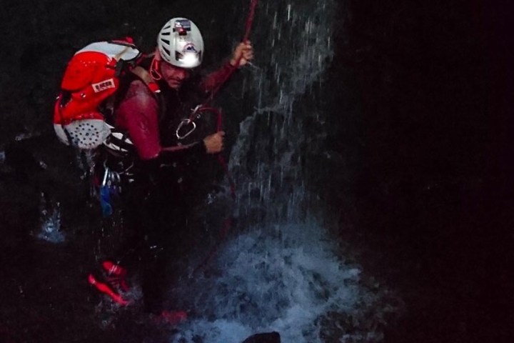 Person rappelling down a waterfall in dark conditions, wearing helmet and gear.