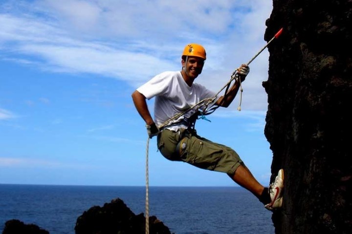 Person rock climbing with a rope and harness, wearing an orange helmet, against a cliff by the ocean.