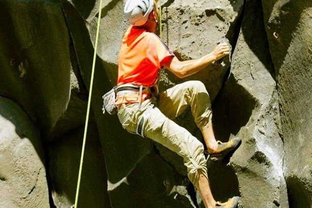 Person rock climbing on a steep face, wearing a helmet and orange shirt.